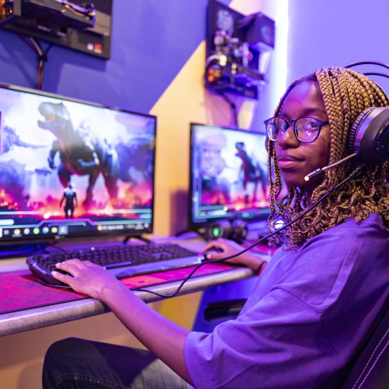 Young woman wearing headset and playing a futuristic online video game fighting a dinosaur using her computer and mouse in a purple gaming room